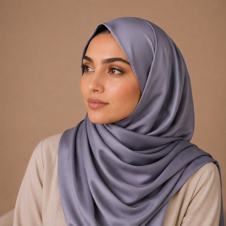 A woman wearing a draped, satin-like dusty purple hijab and a light beige top, set against a solid warm beige background.